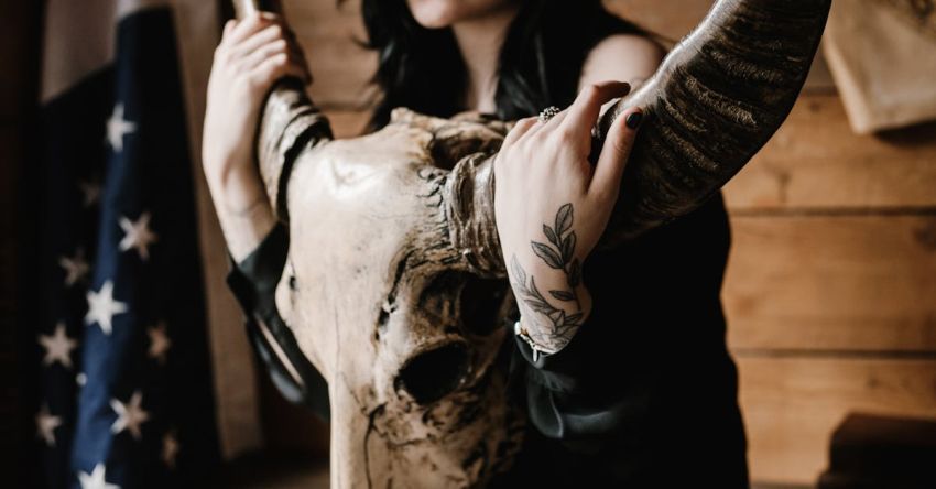 Bold Trends - Crop young brunette with tattooed arms and legs with long horned animal skull sitting near wooden wall with US flag